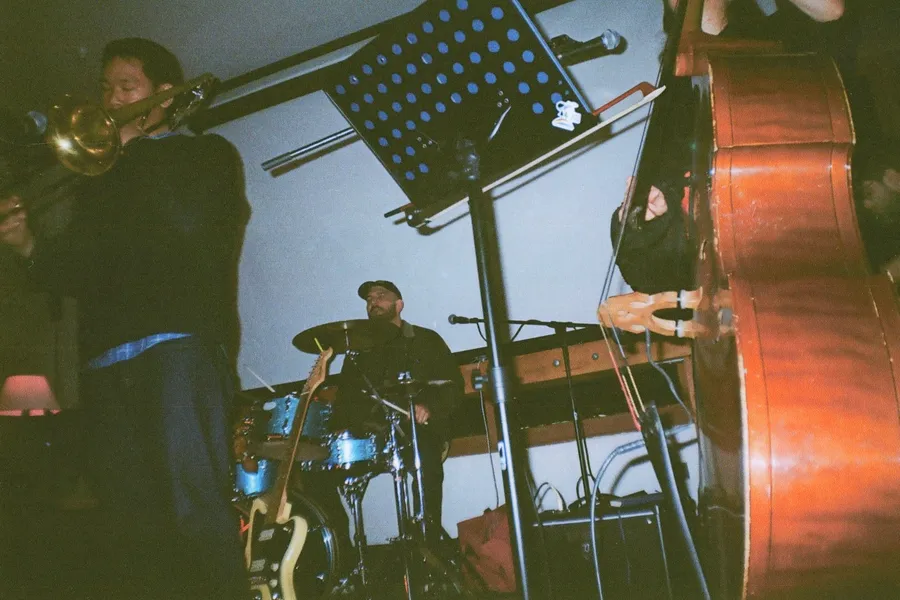 The band mid-set in a dim room — trombone in the foreground, drums behind, upright bass at the right