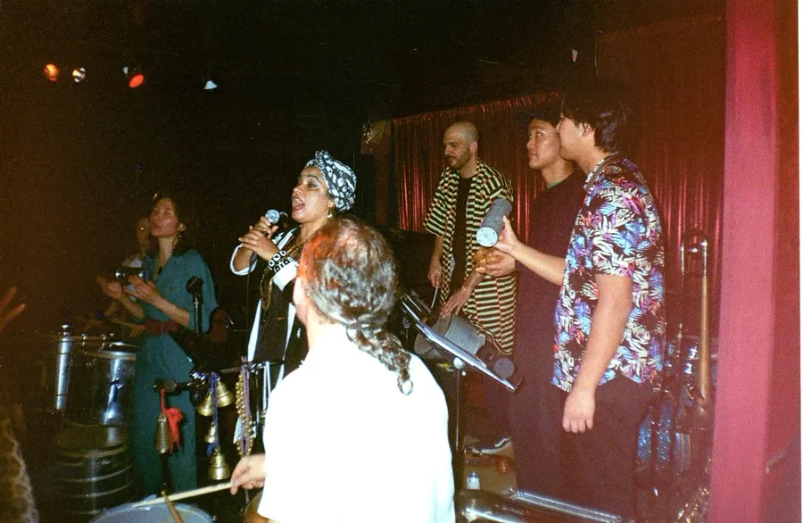 A singer at the microphone in a headwrap, with percussion and horns behind her on a red-curtained stage