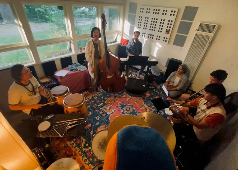 The band rehearsing in a sunlit room, with upright bass, keyboard, congas and horns